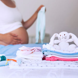 Pile of baby clothes, necessities and pregnant woman on bed in home interior of bedroom.