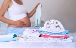 Pile of baby clothes, necessities and pregnant woman on bed in home interior of bedroom.